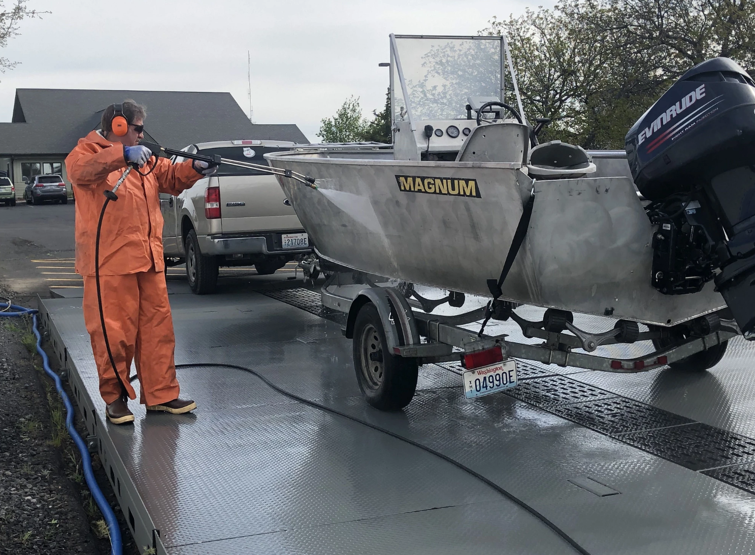 Person in orange protective rain gear, gloves, and ear protection pressure-washing a metal fishing boat on a trailer. The boat, labeled “MAGNUM,” is hit with a spray of water while parked on a wash-down platform beside a pickup truck, with an outboard motor mounted at the stern.